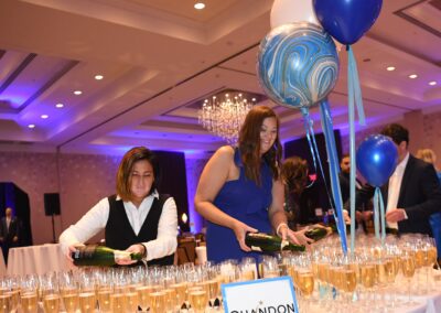 Two women pouring champagne into glasses at an event with blue balloons and a sign reading "chandon.