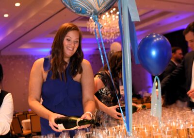 Woman in blue dress serving champagne at an event, holding balloons with a table full of glasses in front.