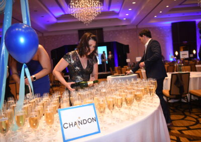A woman pours champagne into glasses at a formal event with a "chandon" sign on the table, set in a ballroom with chandeliers.