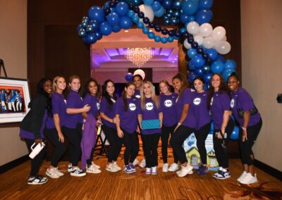 Group of smiling women in purple shirts posing under a balloon arch at an indoor event.