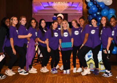 Group of female students in matching purple lsu shirts posing at an event with balloons and a chandelier in the background.