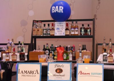 A well-stocked bar with assorted liquor bottles and branded signs for lamarca prosecco, tito's handmade vodka, and beam suntory. a blue balloon floats above.