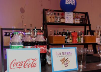 A bar setup with bottles of coca-cola and other beverages, glasses, and brand signs on a table, with a blue "bar" balloon above.