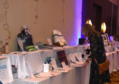 Woman in a floral dress examining items at a silent auction in a banquet hall with various displays on tables.