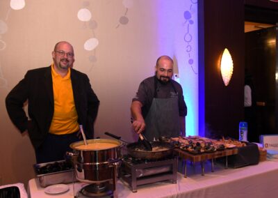 Two men at a catered event, one stirring a skillet and the other standing by a soup pot, with a decorated buffet setup in the background.