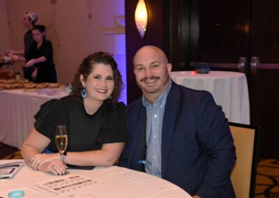 A man and a woman sitting at a table with a champagne glass and event materials, smiling at the camera during a formal gathering.