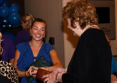 Two women smiling and shaking hands at an indoor event, one young wearing a blue dress and the other elderly with curly hair.