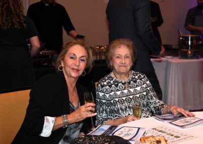 Two elderly women sitting at a banquet table, one holding a glass of wine, with food plates in front of them and people in the background.