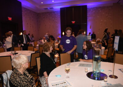 People networking at a banquet hall event, with attendees wearing casual and formal wear, some standing and some seated at tables.