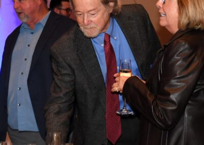 Elderly man with a red tie and a glass of champagne looks at a table filled with champagne glasses, with two people in the background.